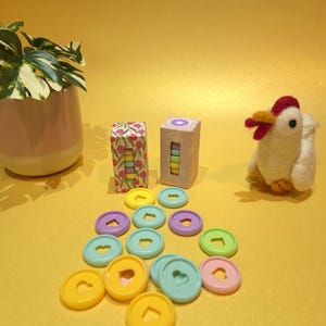 Op de afbeelding: Een set kleurrijke plastic bindclips met hartvormige uitsparingen. De clips zijn in een cirkel op een geel oppervlak gerangschikt. Er zijn twee kleine dozen met kleurrijke ontwerpen op de achtergrond.