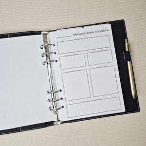 May include: A black six-ring binder containing a white page with the German header W&Ouml;CHENTLICHER R&Uuml;CKBLICK. The page features sections for weekly highlights, successes, improvements, unexpected positives, insights, and future changes. A gold pen is attached.