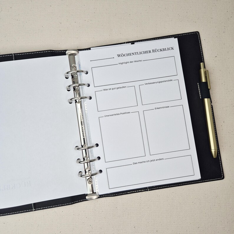 May include: A black six-ring binder containing a white page with the German header W&Ouml;CHENTLICHER R&Uuml;CKBLICK. The page features sections for weekly highlights, successes, improvements, unexpected positives, insights, and future changes. A gold pen is attached.