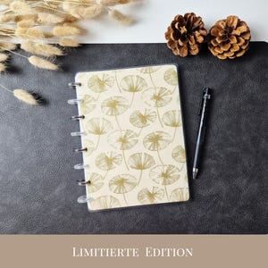 May include: A disc-bound notebook with a cream-colored cover featuring a repeating gold line-art pattern of dandelion seed heads. A black ballpoint pen rests beside the notebook. The text LIMITIERTE EDITION is printed at the bottom.