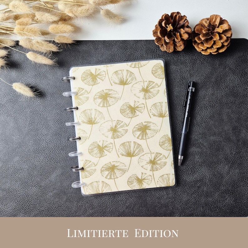 May include: A disc-bound notebook with a cream-colored cover featuring a repeating gold line-art pattern of dandelion seed heads. A black ballpoint pen rests beside the notebook. The text LIMITIERTE EDITION is printed at the bottom.