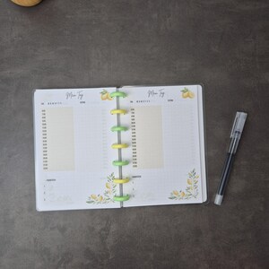 May include: An open planner with a clear cover, featuring a lemon and leaf design. The planner has yellow and green binding rings and a pen. The pages are white with time slots and the German text "Mein Tag".
