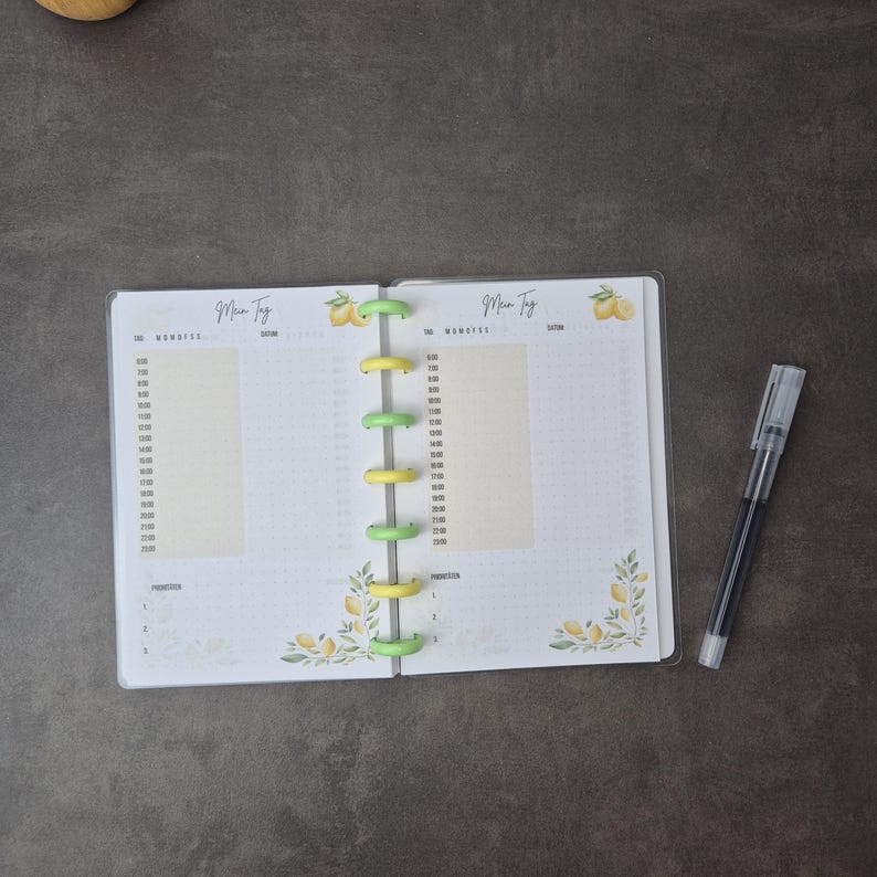 May include: An open planner with a clear cover, featuring a lemon and leaf design. The planner has yellow and green binding rings and a pen. The pages are white with time slots and the German text "Mein Tag".