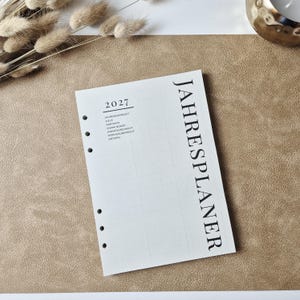 May include: A white planner insert page with six punched holes along the left side. The text 2027 is printed at the top left, followed by a list of sections including Jahres&uuml;bersicht, Ziele, Kontakte, Vision Board, Monats&uuml;bersicht, Wochen&uuml;bersicht, and Notizen. The word JAHRESPLANER is printed vertically in large black letters on the right side.