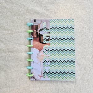 Peut inclure: Un petit carnet avec une couverture à chevrons verts et blancs. Le carnet est maintenu par des anneaux en plastique colorés. Les pages intérieures présentent une photographie d'une cafetière et d'une bougie.