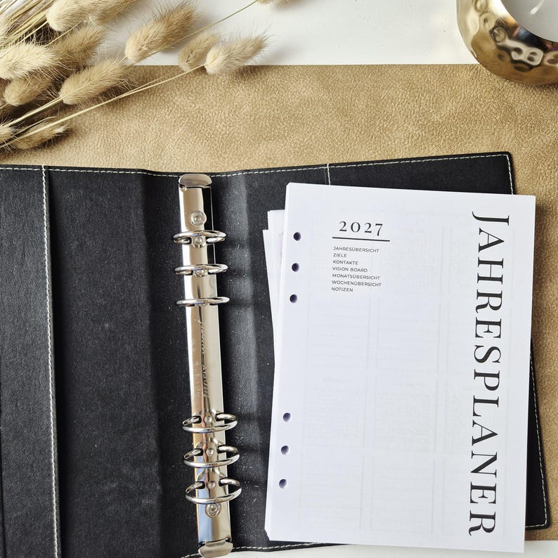 May include: A black six-ring binder with white stitching containing white planner pages. The top page features the year 2027 and the German text JAHRESPLANER, JAHRES&Uuml;BERSICHT, ZIELE, KONTAKTE, VISION BOARD, MONATS&Uuml;BERSICHT, WOCHEN&Uuml;BERSICHT, and NOTIZEN.
