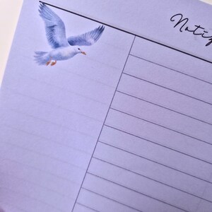 May include: A light purple lined notepad with a watercolor illustration of a seagull in flight. The word "Notiz" is written in cursive in the upper right corner.
