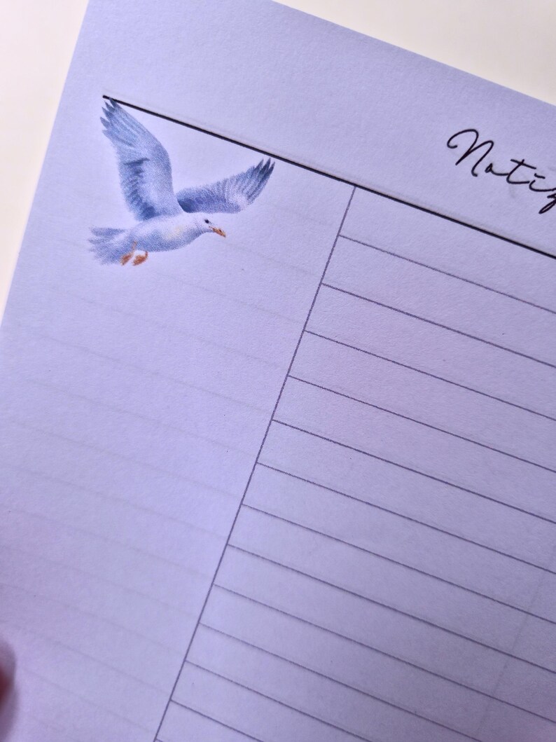 May include: A light purple lined notepad with a watercolor illustration of a seagull in flight. The word "Notiz" is written in cursive in the upper right corner.