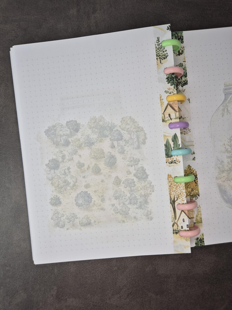 May include: A dot grid journal with a watercolor illustration of a forest scene. The journal has a colorful binding with a forest pattern and multi-colored plastic rings. The pages are white.