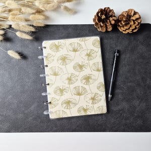 May include: A disc-bound notebook featuring a cream-colored cover with a repeating gold-toned line art pattern of dandelion-like seed heads. The notebook is accompanied by a black ballpoint pen, positioned to the right of the cover.