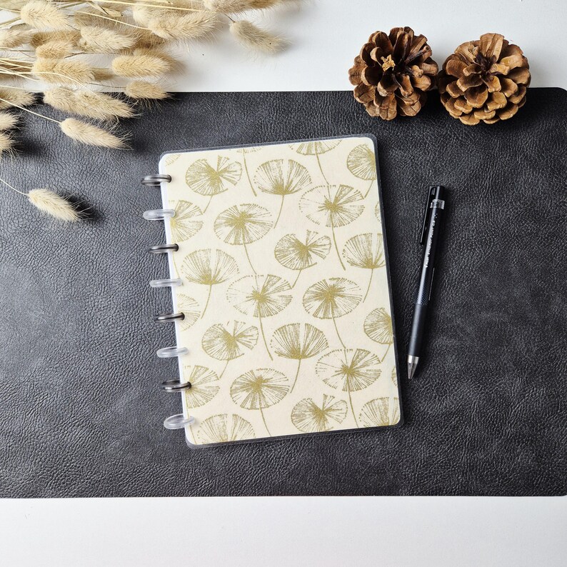 May include: A disc-bound notebook featuring a cream-colored cover with a repeating gold-toned line art pattern of dandelion-like seed heads. The notebook is accompanied by a black ballpoint pen, positioned to the right of the cover.