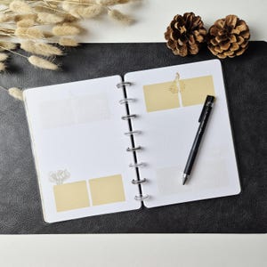 May include: An open disc-bound planner with white dotted pages featuring light yellow rectangular adhesive notes and a black ballpoint pen resting on the right page. The planner includes subtle floral illustrations and is displayed on a black surface.