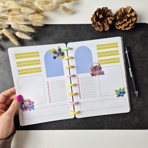 May include: An open disc-bound planner with white pages featuring yellow horizontal bars, light blue arch shapes, and berry illustrations including blackberries and blueberries. The planner has pastel-coloured discs and is accompanied by a black pen.