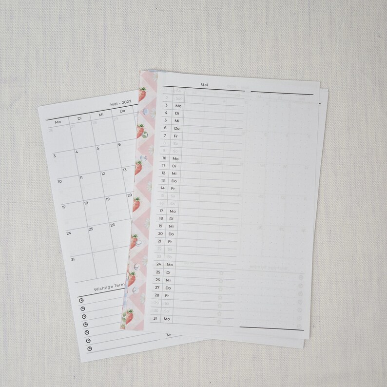 May include: Two white paper calendar pages with the month of May 2027 printed on them. One page is a calendar view, and the other is a planner view. A pink and white strawberry-patterned border is on the left side.