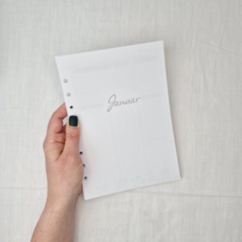 May include: A white planner page with the word "Januar" in cursive script. The page has pre-punched holes for binding and is held by a hand with a dark green nail polish. The background is a light, textured surface.