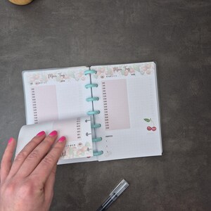 May include: Open planner with a light blue binding, featuring a pink and white cherry design. The pages are divided into time slots and include the German phrase "Mein Tag". A black pen rests on the dark gray surface.