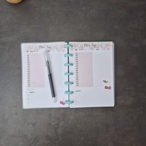 May include: Open planner with a light blue binding and the words "Mein Tag" at the top. The pages have a pink background with time slots and cherry illustrations. A black pen rests on the open pages.