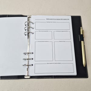 May include: A black leather-like binder with white stitching, featuring a six-ring metal mechanism holding a white page titled W&ouml;chentlicher R&uuml;ckblick. The page includes sections for weekly highlights, successes, improvements, and insights. A gold pen is attached.