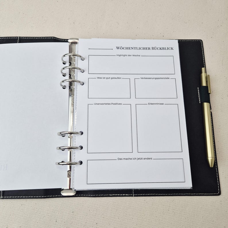 May include: A black leather-like binder with white stitching, featuring a six-ring metal mechanism holding a white page titled W&ouml;chentlicher R&uuml;ckblick. The page includes sections for weekly highlights, successes, improvements, and insights. A gold pen is attached.