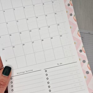 May include: A close-up of a white planner page with a monthly calendar grid and a "To do" list. The page includes the text "Wichtige Termine" and is held by a hand with a dark green nail polish. The planner has a pink floral border.