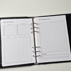 K&ouml;nnte beinhalten: Ein schwarzer Ringbuchplaner, der auf zwei Seiten aufgeschlagen ist. Die linke Seite ist mit ABO TRACKER betitelt und enth&auml;lt Abschnitte f&uuml;r Wunschliste, Kategorien sowie eine Tabelle f&uuml;r Abo, Monat, Jahr und Kosten. Rechts steht ABO &Uuml;BERSICHT &euro;.