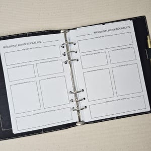May include: A black six-ring binder containing white planner pages with the German text W&ouml;chentlicher R&uuml;ckblick. The pages feature sections for weekly highlights, successes, improvement areas, unexpected positives, insights, and future changes.