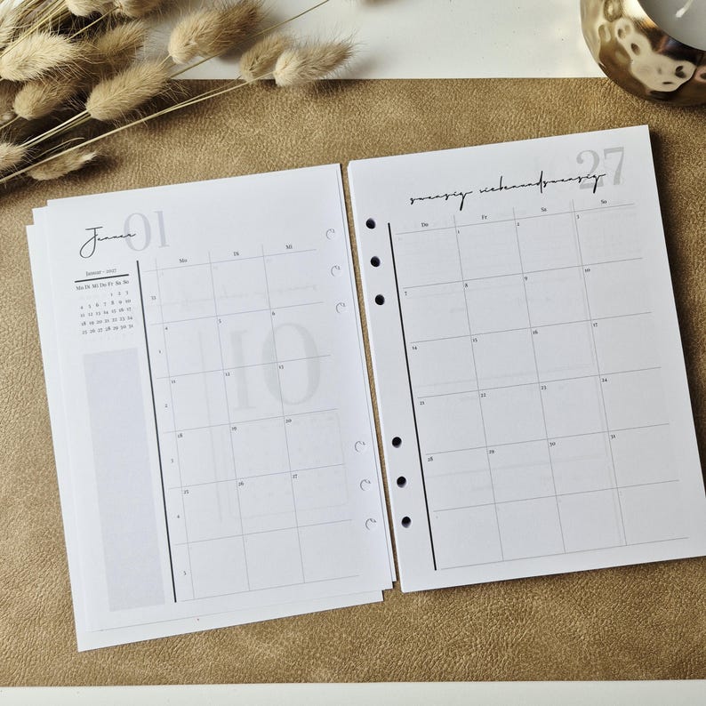 May include: Two white planner pages with black ink. The left page shows a monthly calendar for January with a small overview and a large number 01. The right page features a grid layout for dates with the number 27 in the corner and punch holes.