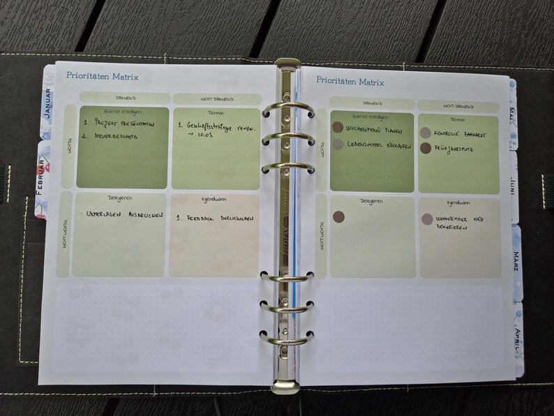 K&ouml;nnte beinhalten: Offener Planer mit dem Titel "Prioritaten Matrix" auf Deutsch. Der Planer ist auf einer Doppelseite mit gr&uuml;nen und beigen K&auml;stchen ge&ouml;ffnet, die mit handschriftlichen Notizen gef&uuml;llt sind. Der Planer ist mit silbernen Ringen gebunden und hat Monatsregister.
