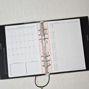 May include: Open black leather planner with a silver binder. The planner is open to a September 2021 calendar and a lined page for notes. A decorative pink divider with strawberry illustrations separates the pages.