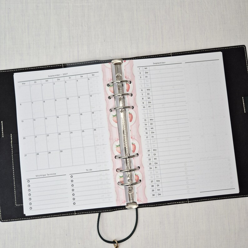 May include: Open black leather planner with a silver binder. The planner is open to a September 2021 calendar and a lined page for notes. A decorative pink divider with strawberry illustrations separates the pages.