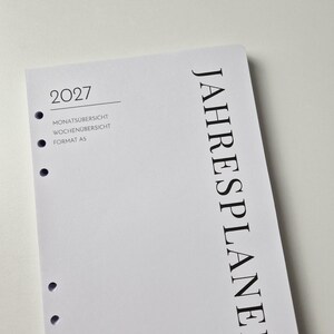 K&ouml;nnte beinhalten: Eine wei&szlig;e A5-Planer-Einlage f&uuml;r das Jahr 2027 mit schwarzem Text. Die Seite zeigt den vertikalen Text JAHRESPLANER, das Jahr 2027 sowie die Begriffe MONATS&Uuml;BERSICHT und WOCHEN&Uuml;BERSICHT. Links befindet sich eine Spalte mit runden Ringbuchlocher.