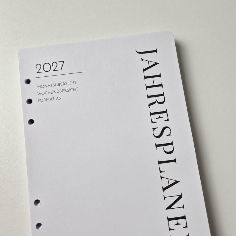 K&ouml;nnte beinhalten: Eine wei&szlig;e A5-Planer-Einlage f&uuml;r das Jahr 2027 mit schwarzem Text. Die Seite zeigt den vertikalen Text JAHRESPLANER, das Jahr 2027 sowie die Begriffe MONATS&Uuml;BERSICHT und WOCHEN&Uuml;BERSICHT. Links befindet sich eine Spalte mit runden Ringbuchlocher.
