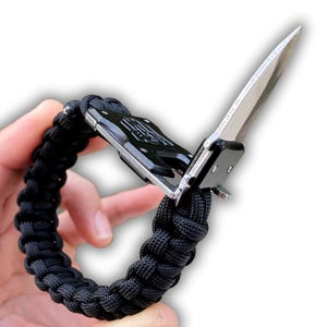 May include: Black paracord bracelet with an integrated folding knife. The knife blade is silver and the handle is black. The bracelet is made of tightly woven black cord. The Paracordgra logo is visible in the bottom left corner.