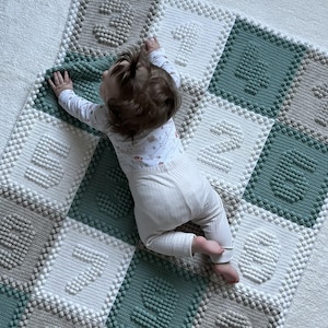 Könnte beinhalten: Ein Baby liegt auf einer handgefertigten, quadratisch gemusterten Decke. Die Decke hat ein sich wiederholendes Muster aus weißen, grauen und teal-farbenen Quadraten mit erhabenen, strukturierten Zahlen. Das Baby trägt einen weißen Strampler und eine Hose.