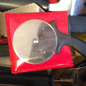 May include: A close-up of a red, black, and silver pizza cutter in a drawer with other kitchen utensils. The pizza cutter is in a red plastic sleeve.