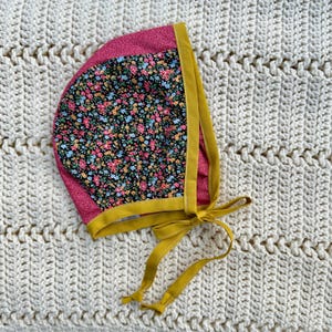 May include: A pink and yellow bonnet with a floral print. The bonnet has a yellow trim and ties.