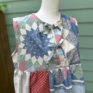 May include: A sleeveless top featuring a patchwork quilt design with various floral and geometric patterns in shades of blue, red, cream, and green. The garment has a round neckline with two fabric ties at the center front and a gathered waistline.