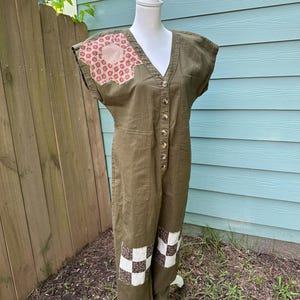 May include: Olive green jumpsuit with a V-neck, button-up front, and cap sleeves. Features decorative patches on the shoulder and knees with floral and checkered patterns. The jumpsuit is displayed on a white mannequin.