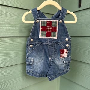 May include: Blue denim baby overalls with a patchwork design on the chest and pocket. The patchwork features red, green, and white squares. The overalls have silver button closures and small pockets.