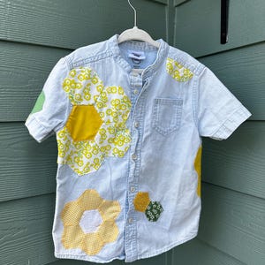 May include: A light blue denim shirt with a patchwork design featuring yellow and green flowers. The shirt has a mandarin collar and short sleeves.