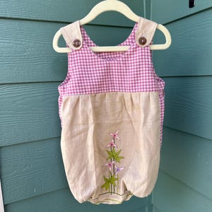 May include: A baby romper with a pink and white gingham top and tan bottom. The romper has tan straps with brown buttons and a floral embroidery design. The romper is hanging on a white hanger.