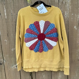 May include: A mustard yellow long-sleeve sweatshirt featuring a large, circular, multi-colored floral appliqué made of quilted fabric segments in red, blue, and white patterns. A white tag attached to the collar reads JOYFULLY MADE Size: M.