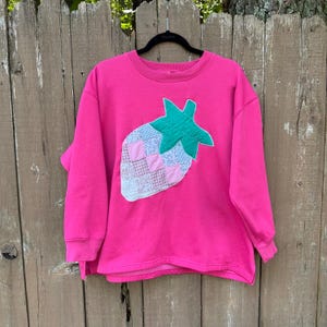 May include: A bright pink long-sleeve sweatshirt featuring a large, central strawberry-shaped appliqué. The strawberry is constructed from a patchwork of various light blue, white, and pink floral and checkered fabric squares with a green leaf top.