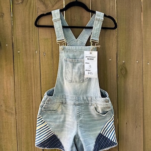 May include: Light blue denim overall shorts with a front pocket and adjustable straps. The shorts have navy blue and white striped accents on the sides. A tag reads "JOYFULLY MADE" with size 3 and a price of $45.