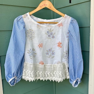 May include: A white blouse with blue gingham sleeves and floral embroidery. The blouse features a lace trim at the bottom and is hanging on a wooden hanger. The sleeves are long and gathered at the cuffs.