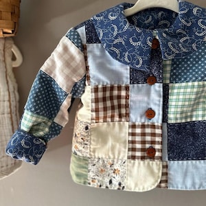 May include: A patchwork jacket in various shades of blue, brown, green, and cream. The jacket features a rounded collar and three brown buttons down the front. The sleeves and collar have a blue floral pattern. The jacket is hanging on a white hanger.