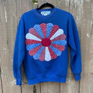 May include: A royal blue long-sleeve sweatshirt featuring a large, circular, multi-colored fabric flower appliqué on the chest. The flower is composed of alternating red, white, and blue patterned fabric petals surrounding a solid red center.