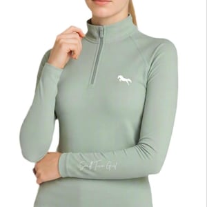 Op de afbeelding: Een saliegroene longsleeve met een kwartritssluiting, voorzien van een wit paard-silhouetlogo op de linkerborst en de tekst Small Town Girl in wit handschrift op de rechteronderarm, gecombineerd met een donkergrijze legging.