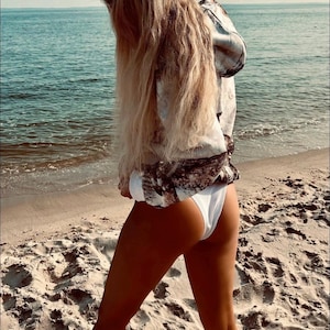 Può includere: Una donna indossa una felpa con cappuccio a motivi bianchi e marroni e slip bikini bianchi in piedi su una spiaggia sabbiosa. Il mare è sullo sfondo.