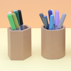 May include: Three brown pen holders with colorful markers. The holders are a hexagon, a cylinder, and a square. A hand is reaching into the square holder to grab a marker. The markers are various colors.